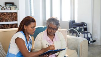 Doctor explaining medication on clipboard to senior woman in living room at home